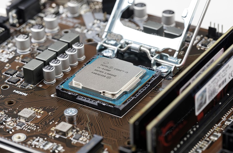 Close-up of a computer motherboard with an Intel Core i5 processor in a socket, surrounded by capacitors and heat sinks. The tone is technical and precise.
